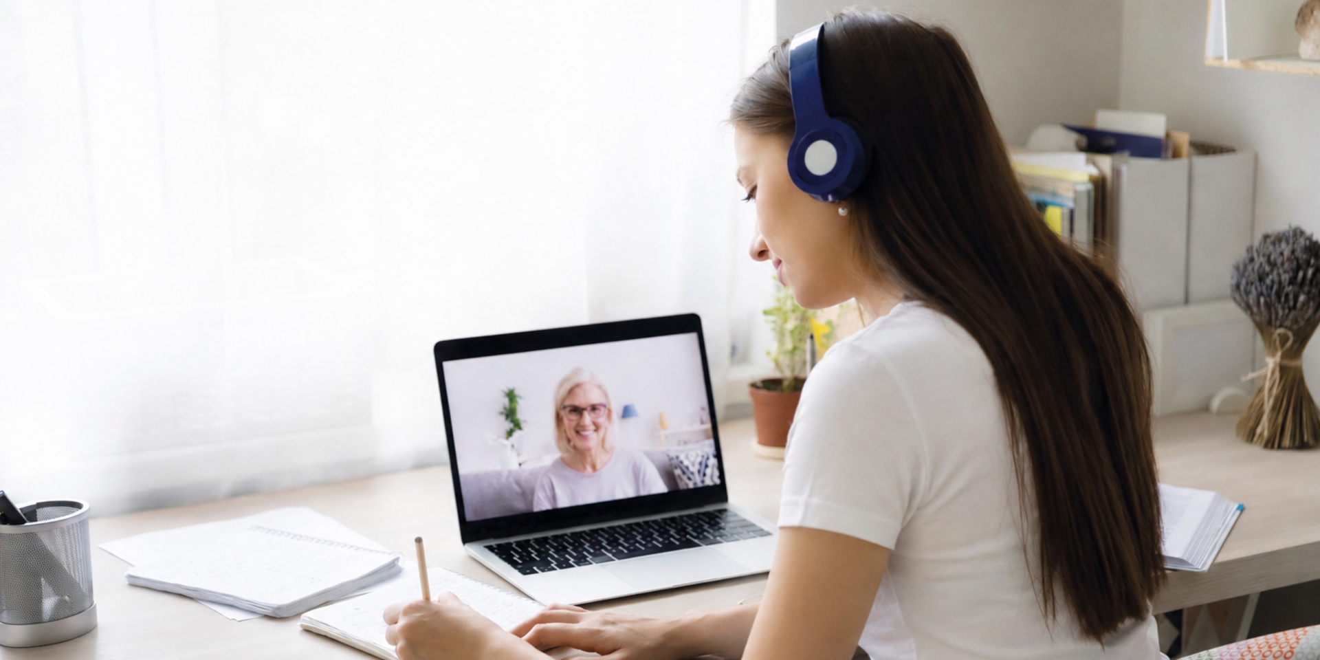 MVZ Online-Psychotherapie | Frau mit langen schwarzen Haaren und weißem Shirt sitzt an einem Tisch. Vor ihr steht ein Laptop und sie macht sich Notizen. Dabei hat sie Kopfhörer auf.